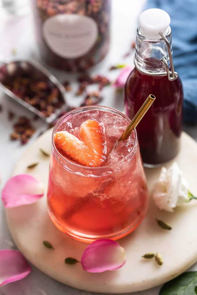 A cardamom rose mocktail in an low ball glass with a heart cut in the shape of a strawberry; rose petals and cardamom pods surround the glass