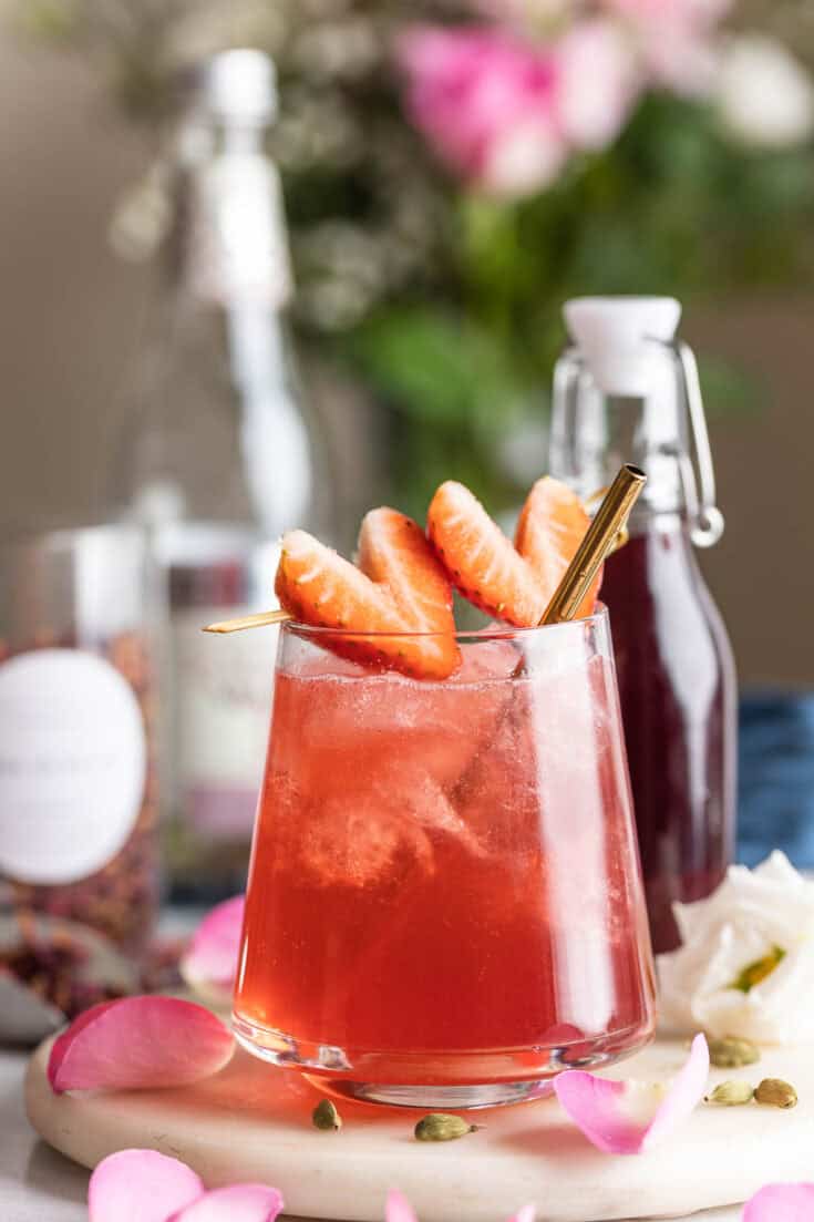 A cardamom rose mocktail in an low ball glass garnished with 2 strawberries cut into the shape of a heart with a heart on a toothpick.