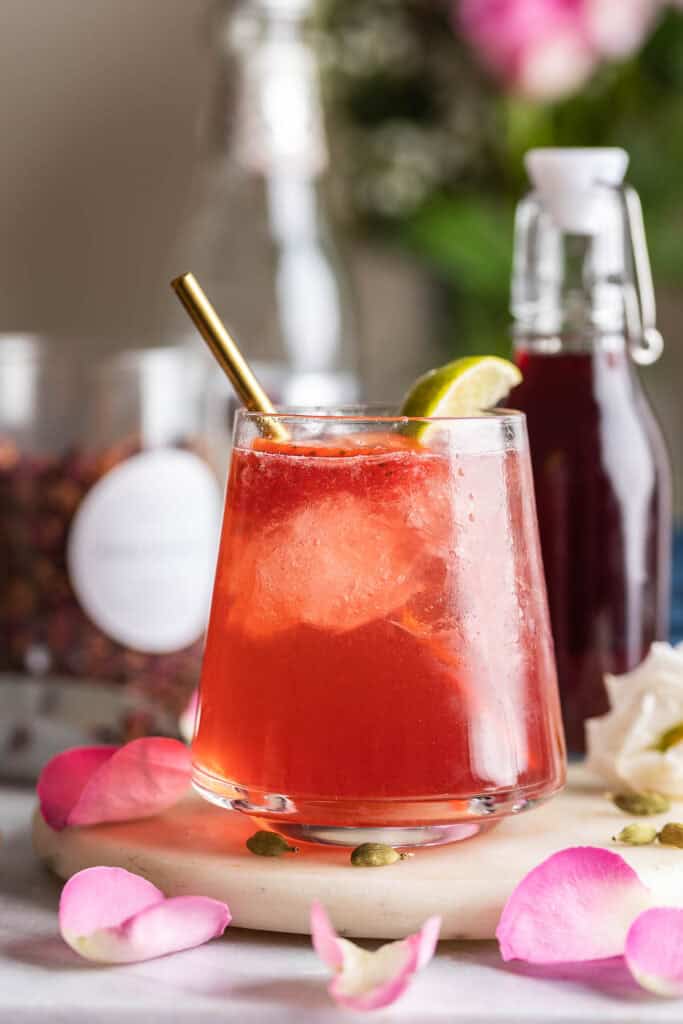 A cardamom rose mocktail in a low ball glass garnished with a lime wedge surrounded by fresh rose petals and cardamom pods.