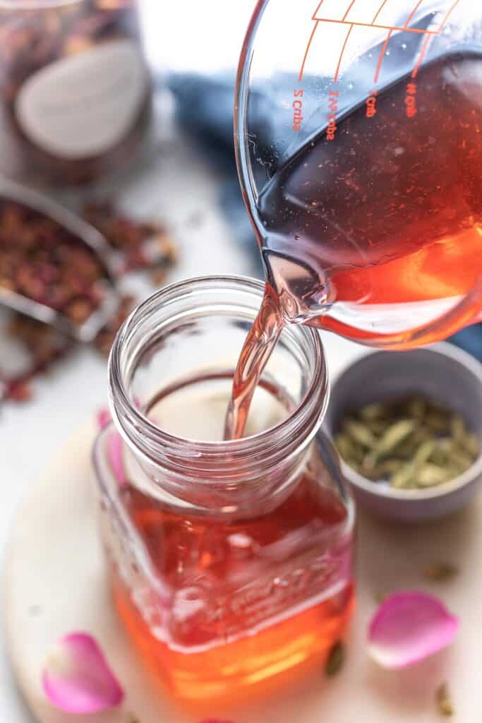 Cardamom Rose Simple Syrup pouring from a measuring jug into a storage jar.