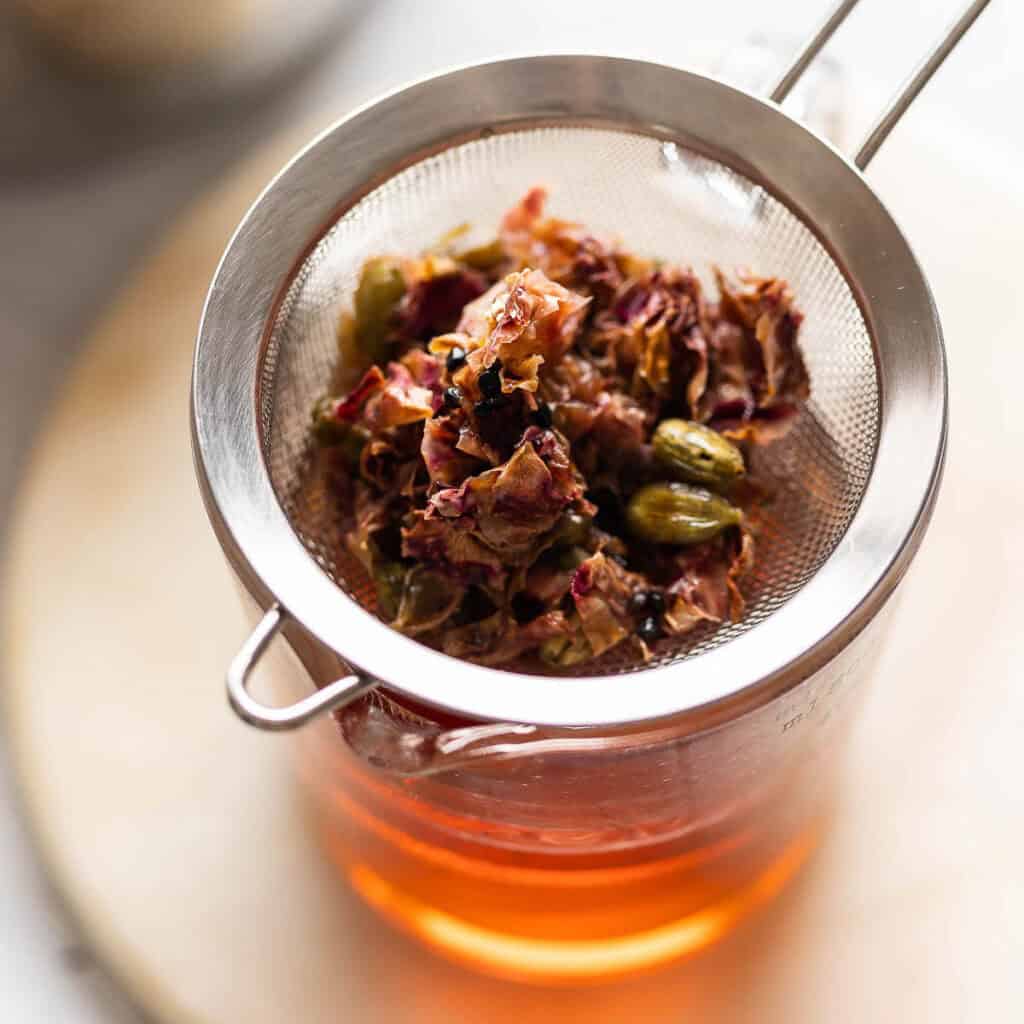 Rose petals and cardamom pods in a fine mesh strainer over a measuring jug full of rose syrup.