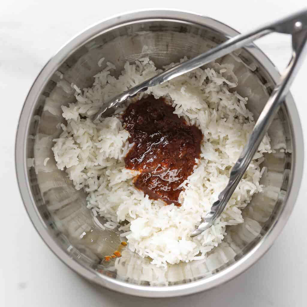 Cooked rice in a bowl with a red curry paste mixture and a pair of cooking tongs to mix it all together.