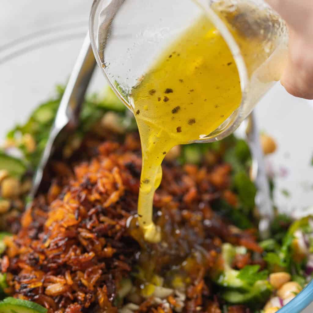 A lemon vinaigrette pouring over a salad with baked rice seasoned with red curry paste, cucumbers, chickpeas and fresh herbs.