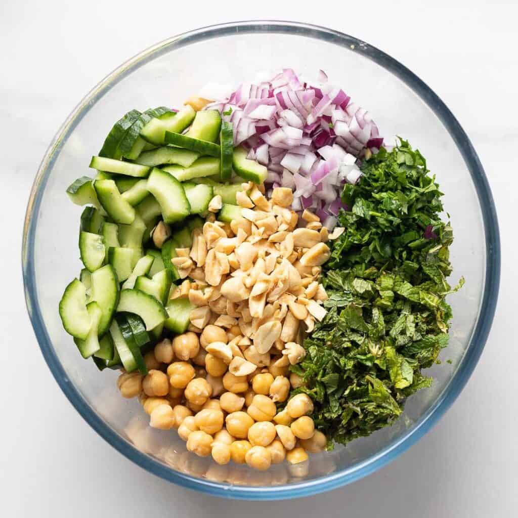 A mixing bowl with diced cucumber, red onion, chopped peanuts, parsley, mint and chickpeas.