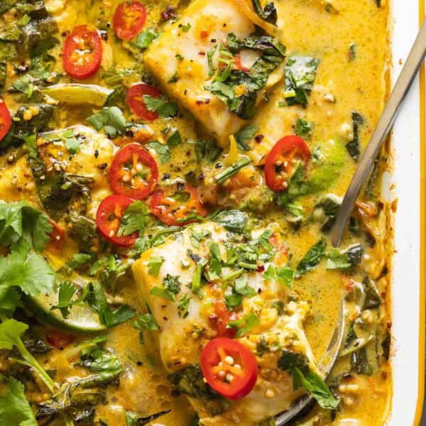 A roasting pan full of A Baked Coconut Fish Curry topped with fresh sliced chilies, cilantro and a serving spoon in side scooping up a piece of white fish covered in curry.