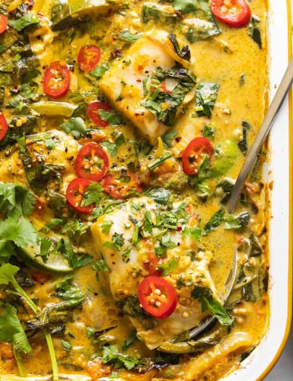 A roasting pan full of A Baked Coconut Fish Curry topped with fresh sliced chilies, cilantro and a serving spoon in side scooping up a piece of white fish covered in curry.