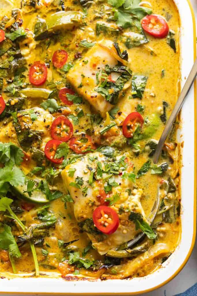 A roasting pan full of A Baked Coconut Fish Curry topped with fresh sliced chilies, cilantro and a serving spoon inside scooping up a piece of white fish covered in curry.