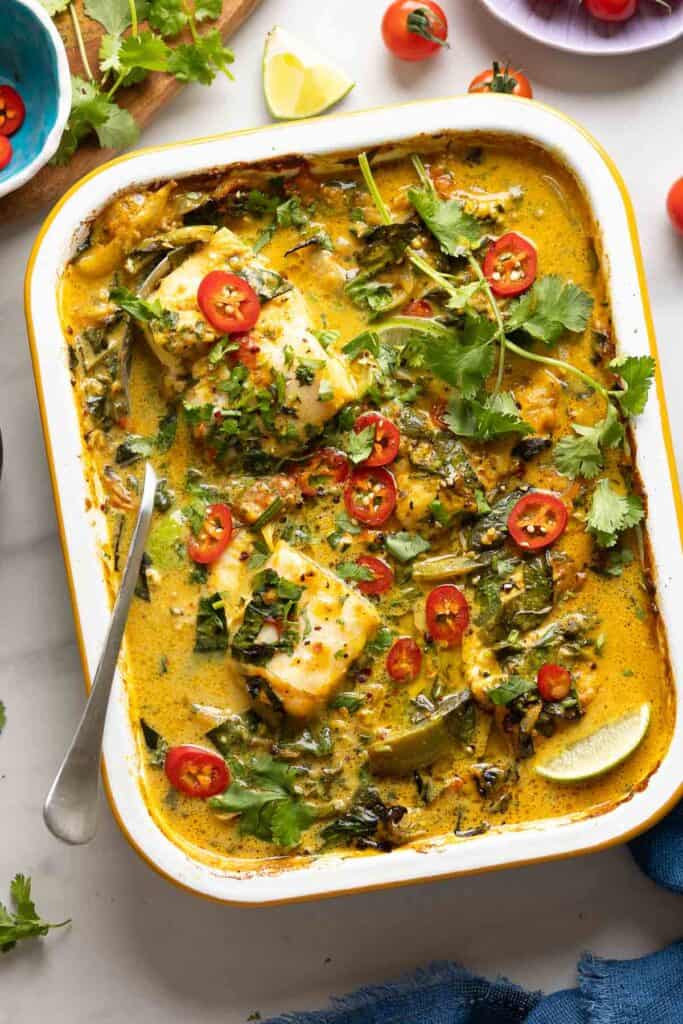 A roasting pan full of A Baked Coconut Fish Curry topped with fresh sliced chilies, cilantro and a serving spoon inside. Fresh tomatoes, lime wedges and cilantro on the side.