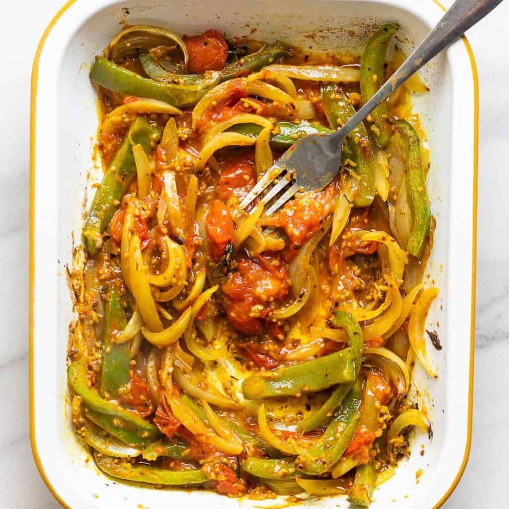 Spiced, roasted tomatoes, onion and bell peppers in a roasting pan with a fork smashing the tomatoes down.