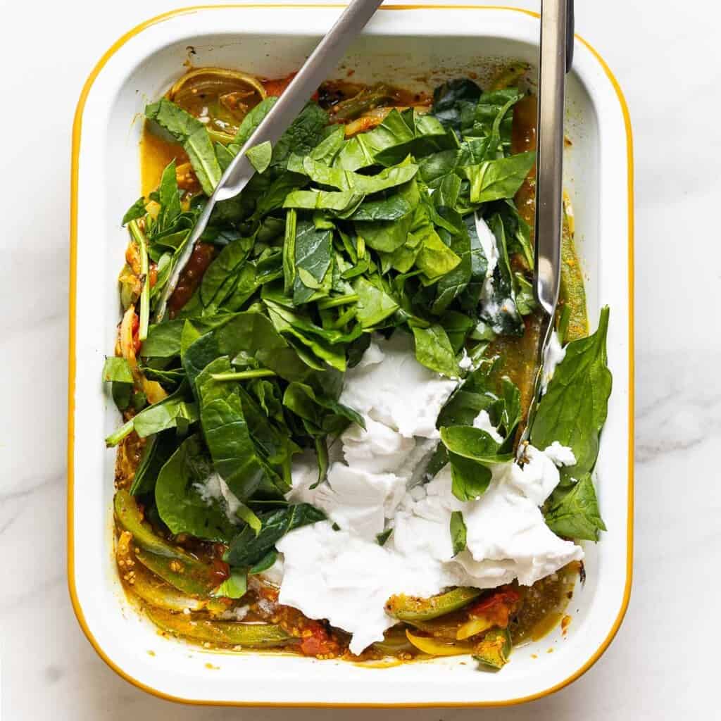 Raw spinach and a can of coconut milk added to a roasting pan full of roasted veggies and a pair of kitchen tongs for mixing.