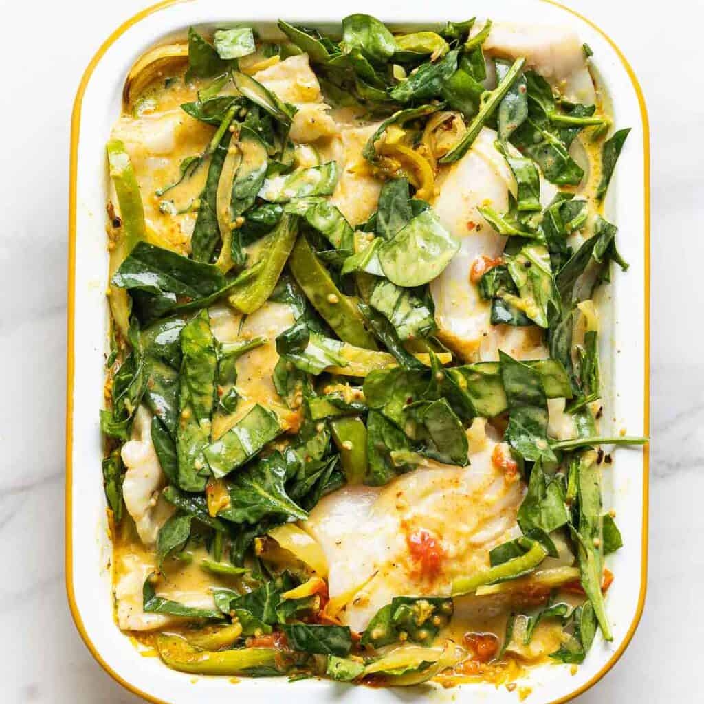 A roasting pan full of roasted vegetables mixed together with raw spinach and coconut milk with pieces of boneless, skinless cod nestled into the spinach.