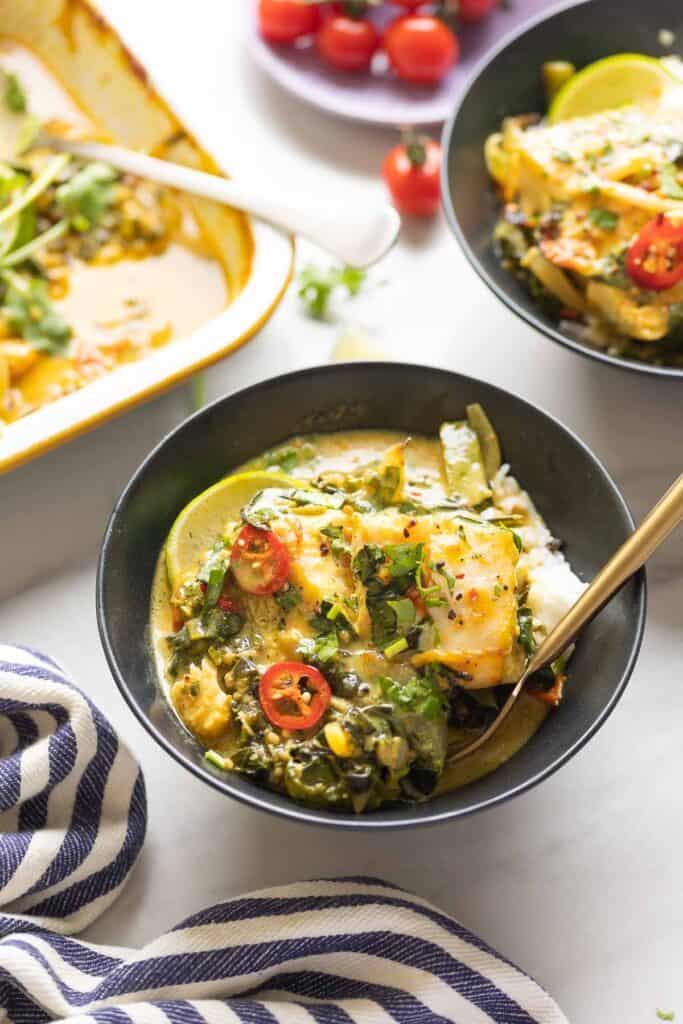 A couple of bowls full of a baked coconut fish curry served over white rice with fresh sliced chilies on top and cherry tomatoes in the back