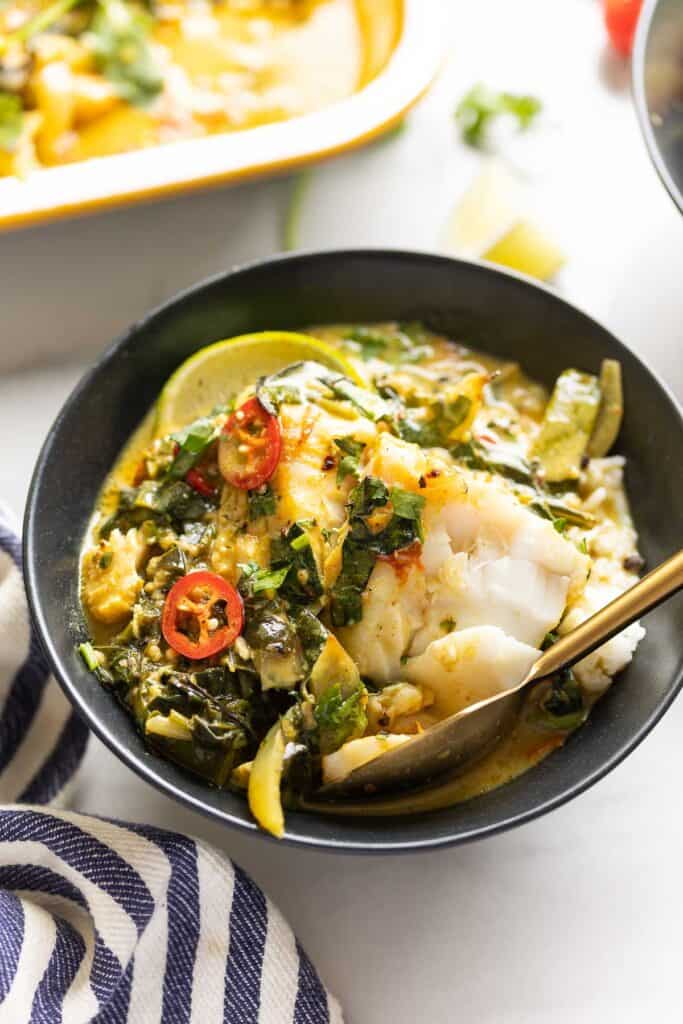 A bowl full of a baked coconut fish curry served over white rice with fresh sliced chilies on top and a spoon cutting into the cod.