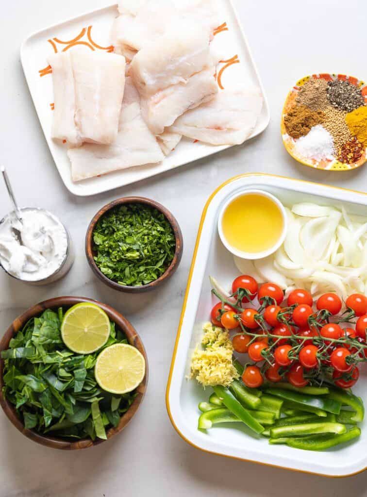 The raw Baked Fish Curry ingredients: boneless, skinless cod, coconut milk in a can, chopped cilantro and spinach, a lime cut in half, sliced onions and bell pepper, grated ginger, cherry tomatoes on the vine, oil and a small plate of spices.