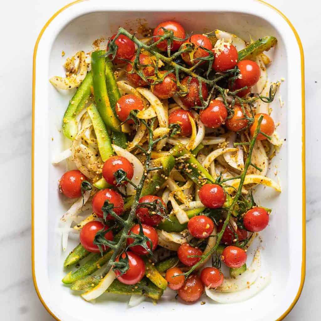 Sliced onions, bell pepper and cherry tomatoes on the vine in a roasting pan tossed in oil and spices.