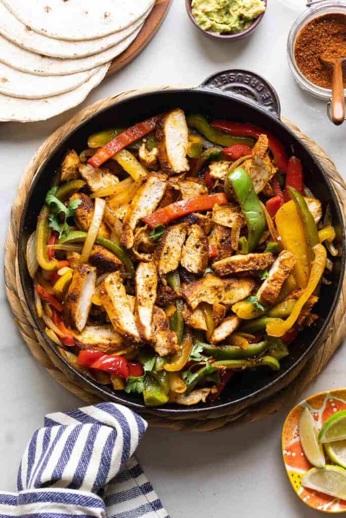 Homemade Chicken Fajitas in a cast iron skillet: sliced chicken breast, sautéd bell peppers and onions mixed together. On the side: flour tortillas, a bowl of guacamole, a small plate of lime wedges and a jar of homemade fajita seasoning.