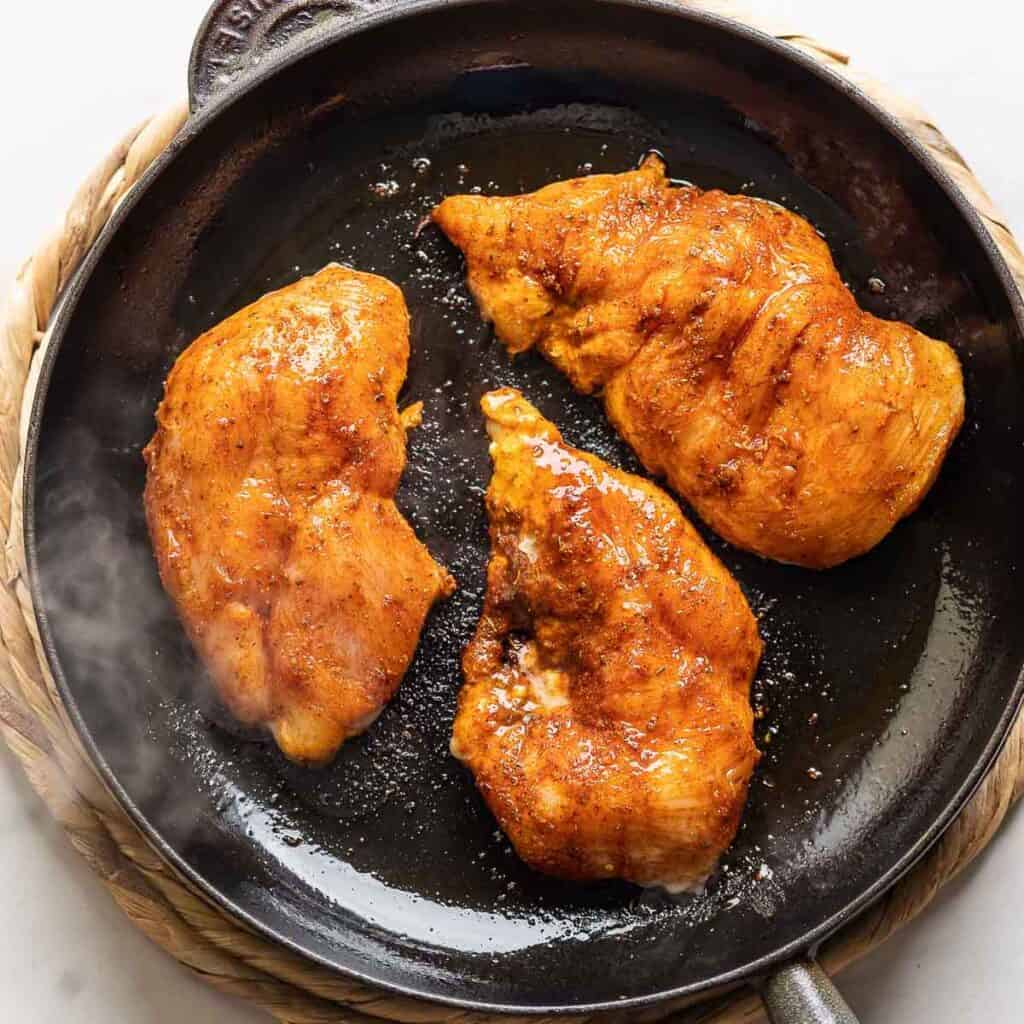 Three chicken breasts coated in a fajita marinade cooking in a cast iron skillet. 