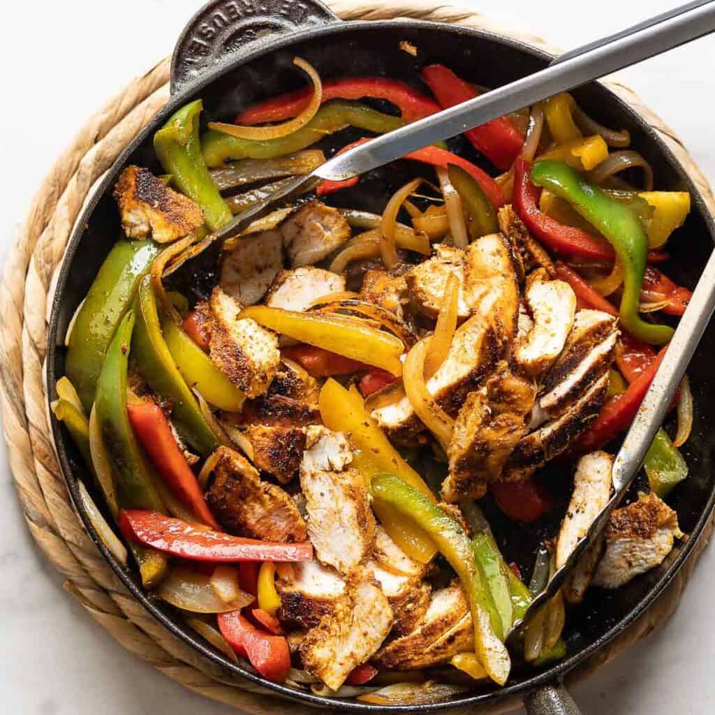 A cast iron skillet chicken fajitas: sliced chicken, sautéd bell peppers and onions mixed together with a pair of tongs.