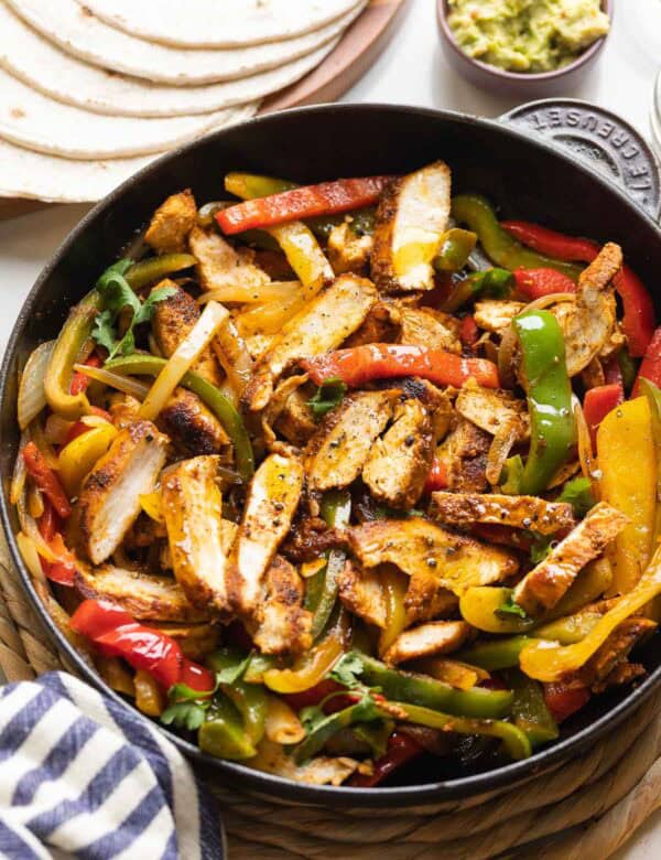 Homemade Chicken Fajitas in a cast iron skillet: sliced chicken breast, sautéd bell peppers and onions mixed together with flour tortillas and guacamole on the side.