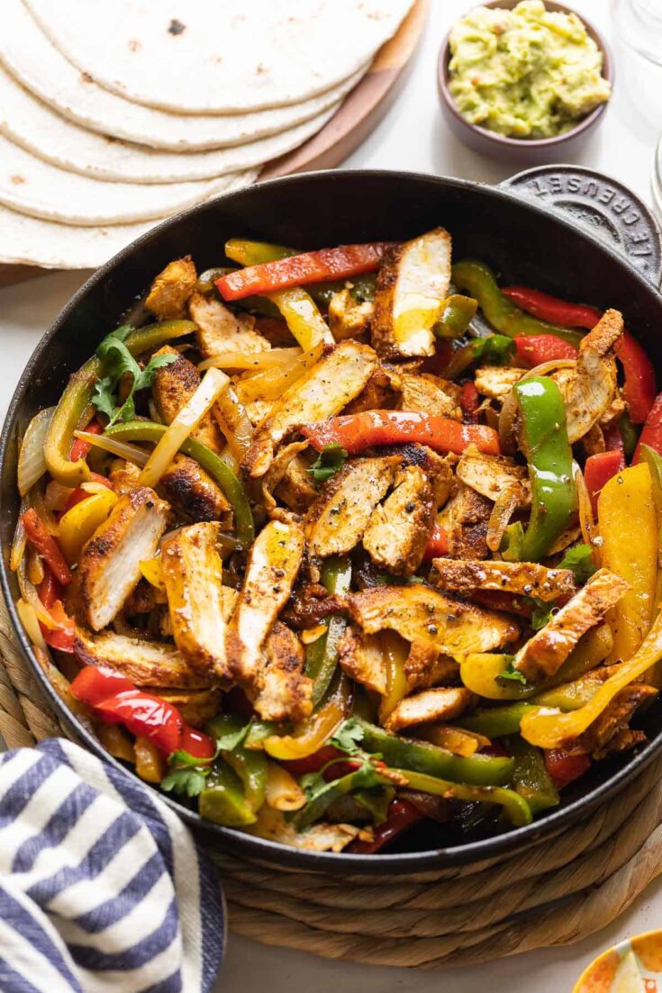 Homemade Chicken Fajitas in a cast iron skillet: sliced chicken breast, sautéd bell peppers and onions mixed together with flour tortillas and guacamole on the side.