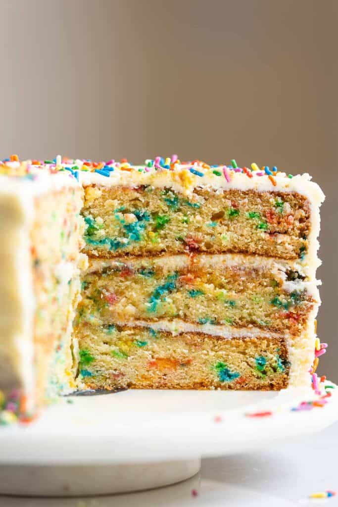 A three layer gluten free funfetti cake decorated with a vanilla frosting and rainbow sprinkles on a cake platter.