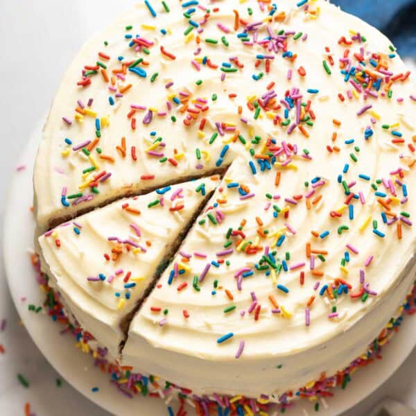 A Gluten Free Funfetti Cake frosted with vanilla buttercream and rainbow sprinkles sitting on a cake platter with a single slice cut into the cake.