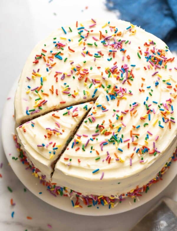 A Gluten Free Funfetti Cake frosted with vanilla buttercream and rainbow sprinkles sitting on a cake platter with a single slice cut into the cake.