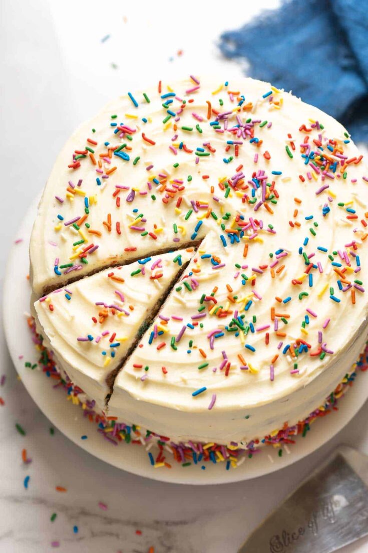 A Gluten Free Funfetti Cake frosted with vanilla buttercream and rainbow sprinkles sitting on a cake platter with a single slice cut into the cake.