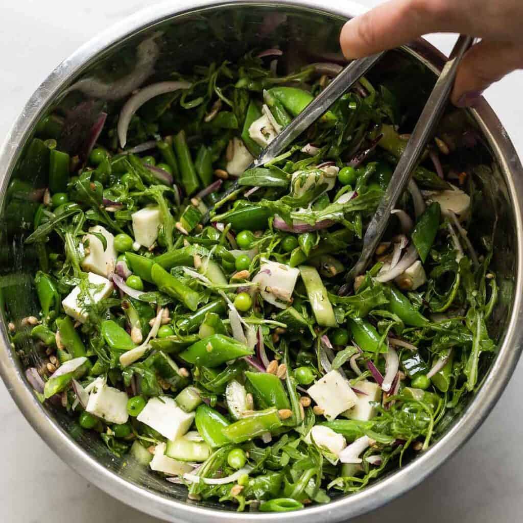 A large salad bowl full of a Spring Greens Salad made up of blanched veggies, arugula, mozzarella, sunflower seeds, onion and cucumber.