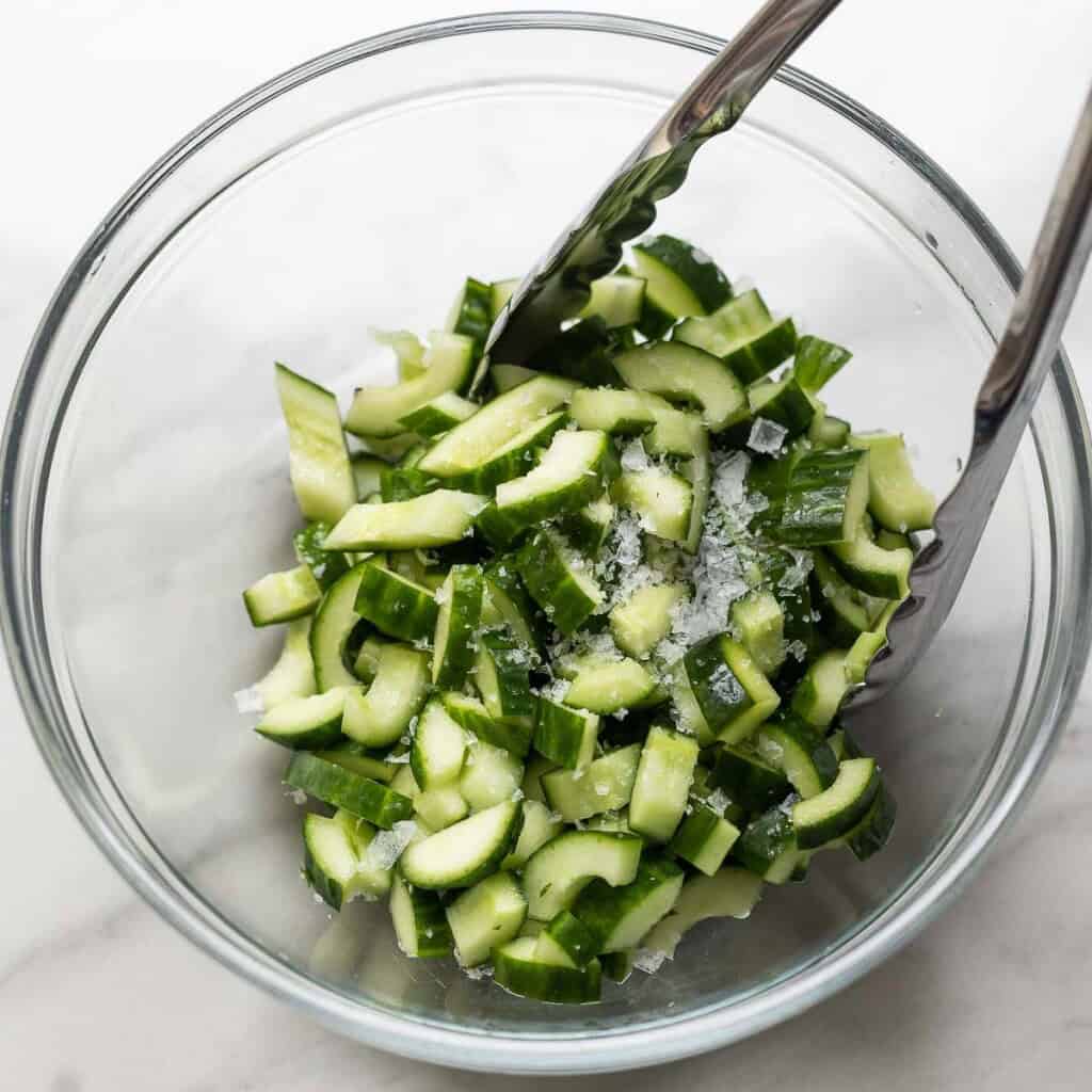 a pair of tongs tossing together diced and de-seeded cucumber with salt