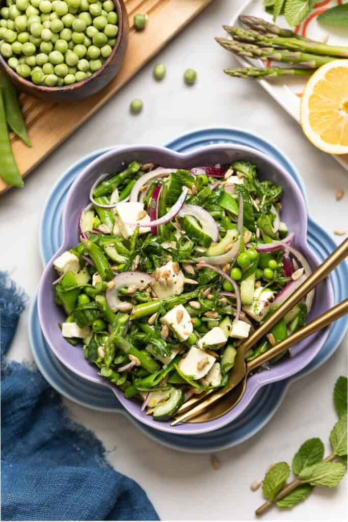 A salad bowl full of a Spring Greens Salad with some of the fresh ingredients surrounding the bowl including: mint, lemon, asparagus, peas and sunflower seeds. 