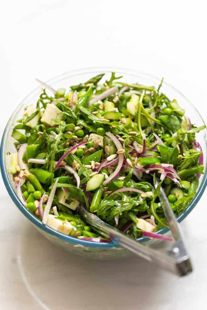 A large mixing bowl full of a Crunchy Spring Greens Salad with a pair of tongs inside the bowl to toss the salad together.