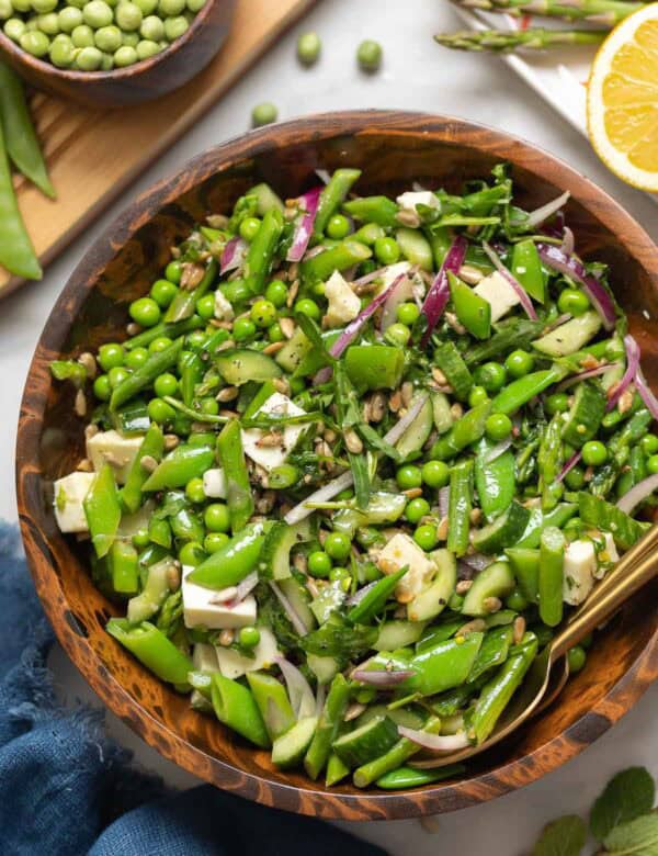 A Spring Greens Salad in a bowl made with snap peas, english peas, asparagus, cucumber, onion, mozzarella and sunflower seeds with the fresh ingredients surrounding the bowl.