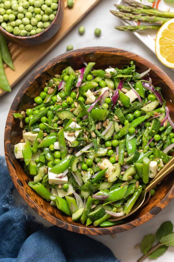 A Spring Greens Salad in a bowl made with snap peas, english peas, asparagus, cucumber, onion, mozzarella and sunflower seeds with the fresh ingredients surrounding the bowl.