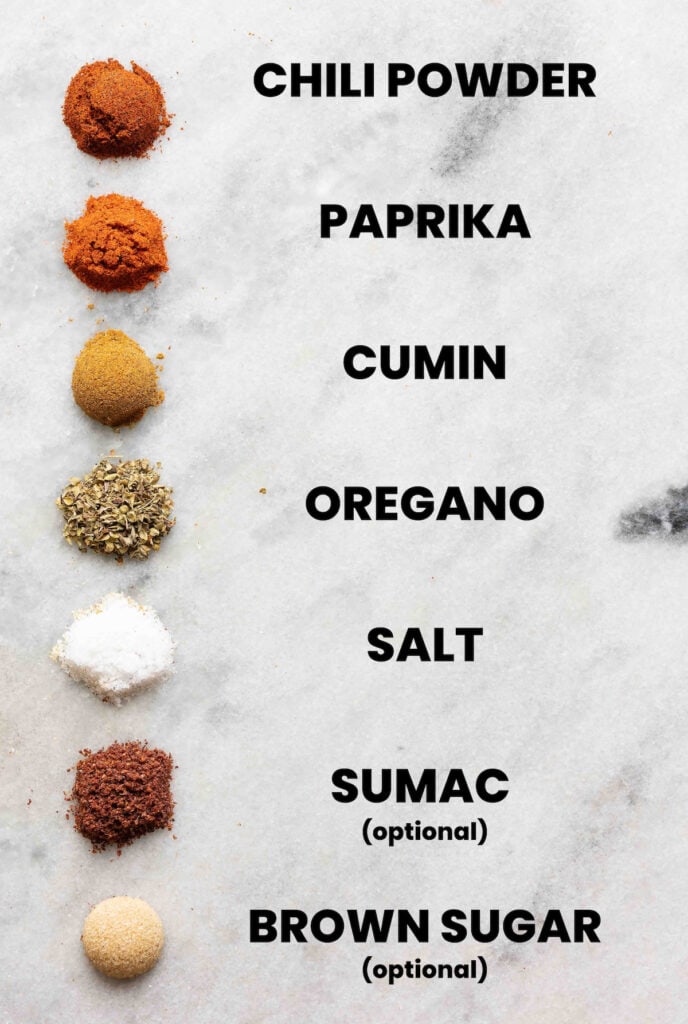 The individual spices needed to make a homemade low fodmap fajita seasoning blend lined up in a row with names listed next to each spice: chili powder, paprika, cumin, oregano, salt, sumac (optional) and brown sugar (optional)